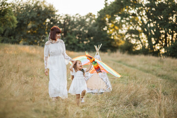 Naklejka na ściany i meble Cute little girl runs with a kite in the summer field during sunset, holds hand of her mother. Happy child enjoying walk with mom, playing with a kite outdoors. Mom and daughter dressed in boho style