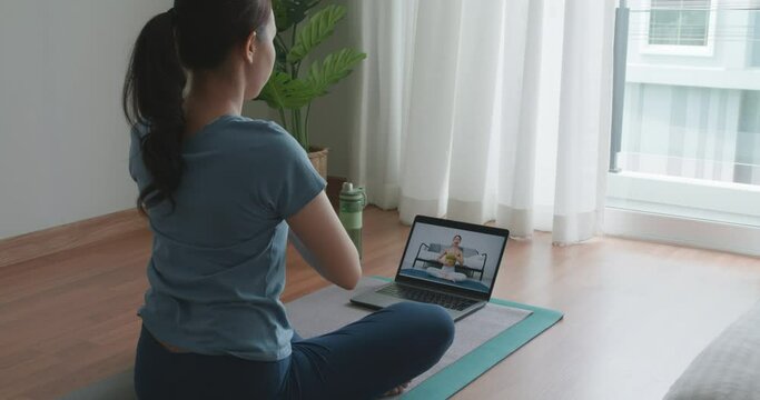Asia people sport woman enjoy study happy watch live video sit on yoga mat at cozy home floor indoor easy zen class. Body calm life stress relax asana lotus pose in laptop self learn online media.