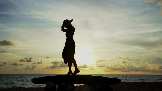 Side view of short hair Asian woman silhouette in a beach dress and straw hat enjoy balancing walking and jump on a beach rocks with a calm sea and the evening sun in beautiful sky in the background.