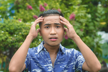 A panicked young Filipino man in a hawaiian shirt. Hands on forehead in shock. Outdoor garden scene.