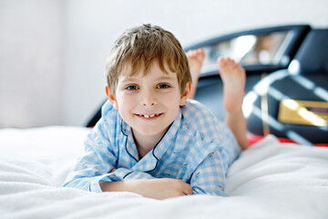 Adorable happy little kid boy after sleeping in his white bed in colorful nightwear. School child celebrating pajama party and looking at the camera. Funny happy child playing and smiling