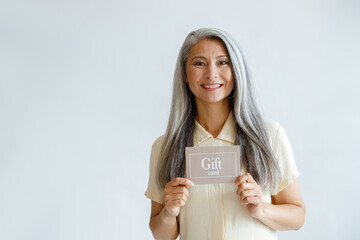 Smiling grey haired Asian woman holds gift card standing on light background in studio, space for...