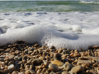 Sea foam surf and shiny multi-colored pebbles. The sea wave has foamed and covers the pebble beach. pebble stones on the sea beach, the rolling waves of the sea with foam