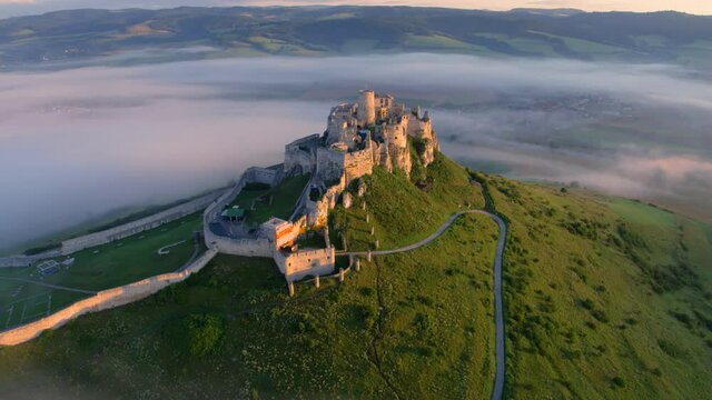 Aerial view of Spi&scaron; Castle, fog flows around the castle, lit by morning sun. Extensive castle ruins, standing on a 200m high white rock above the valley against mountains in background.  UNESCO site.