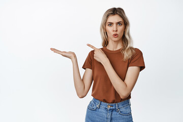 Young woman looks curious while shows smth on her open hand, pointing finger at her palm with product, standing in casual outfit against white background