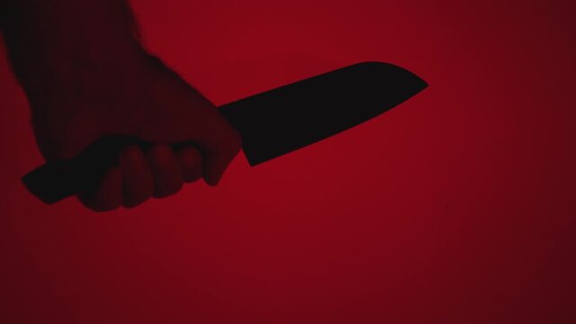 A man's hand holds a large kitchen knife against a red background. The blade of the knife glistens in the light. The concept of a homicidal maniac