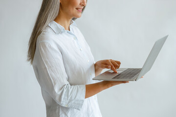 Cheerful hoary haired middle aged lady in white blouse uses modern laptop on light background in...