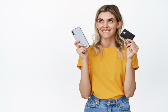 Image of a girl thinking of buying, ordering something from online shop, holding credit card and mobile phone, looking aside thoughtful, white background
