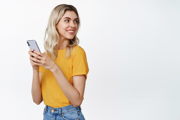 Celluar technology. Young blond woman holds mobile phone, turn head behind and smile at copy space, stands in yellow tshirt over white background