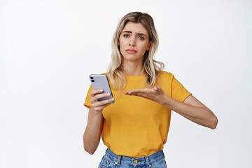 Troubled young woman asking help with mobile phone, pointing at smartphone and looking upset, standing over white background