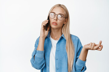 Image of thoughtful woman in glasses talking on mobile phone and shrugging, looking puzzled during...