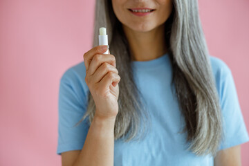 Grey haired woman in blue t-shirt on pink background in studio closeup, focus on hand with open lip balm. Mature beauty lifestyle