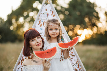Close up of happy family having picnic in summer field. Pretty stylish boho hippie mother with little daughter sitting near wigwam tent and eating watermelon outdoors © sofiko14