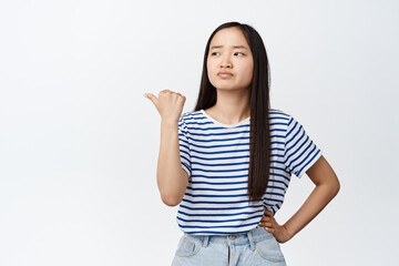 Fototapeta premium Skeptical asian girl pointing and looking left with doubtful face expression, standing in tshirt and jeans over white background