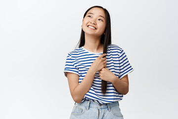 Beautiful asian girl touching her long hair and smiling, dreaming of something, looking up as if picturing moment, standing over white background