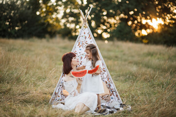 Summer outdoor shot of lovely family, mother and little daughter, spending their picnic time in the wild field, sitting near wigwam tent with watermelon pieces and looking each other with tenderness © Sofiia Shunkina