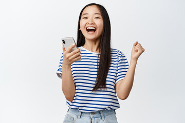 Fototapeta premium Excited asian woman rejoicing from smartphone app notification, winning on mobile phone, standing over white background
