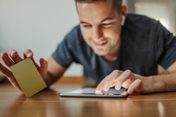 Online purchase with a gold credit card on a tablet. An excited man lies on his stomach on the floor with underfloor heating and uses a tablet with a touchscreen and holds a card in the other hand