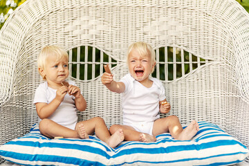 Toddler twins on a canvas chair with pillows Cute babies with blue eyes and blonde hair in white...