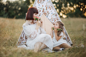 Family, leisure and ethnic people concept. Boho hippie happy mother and little daughter having picnic on summer field, sitting in front of teepee tent and eating watermelon © sofiko14