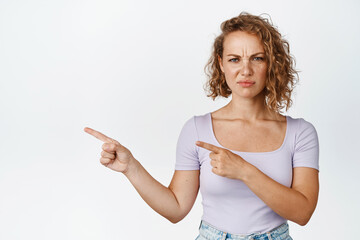 Young blond woman pointing fingers left, frowning and looking with disgust or dismay, complaining,...