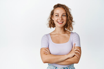 Beautiful blond female model with short curly hairstyle, looking left at copy space and smiling, cross arms on chest, standing in t-shrit over white background