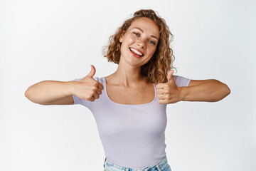 Happy blond woman shows thumbs up and smiles pleased, praise something good, making compliment,...