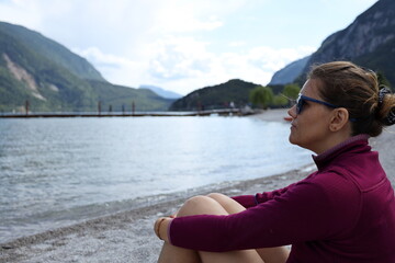 Beautiful girl observes the panorama sitting on the beach of Molveno lake in Trentino, Italy.