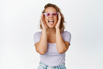 Naklejka premium Wild and free blond girl having fun, wearing sunglasses, showing tongue and shouting carefree, standing happy against white background
