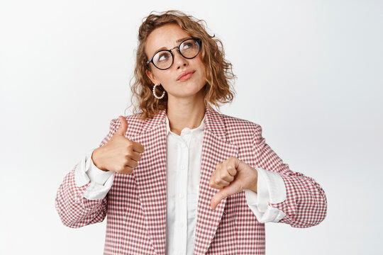Thoughtful businesswoman in glasses and suit, showing thumbs up and down, thinking about something, making decision, weighing pros and cons, white background
