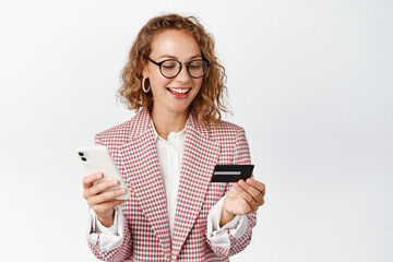 Online shopping. Young female entrepreneut paying on smartphone app, holding mobile phone and credit card, standing against white background