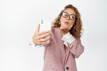 Cute female manager taking selfie, sending kisses at mobile phone camera, romatnic video call on smarpthone app, standing against white background