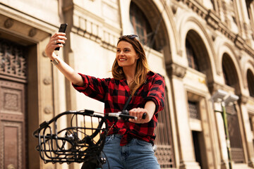 Young blonde woman on bicycle on the street. Beautiful girl taking selfie photo.