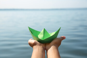 Child holding green paper boat near river, closeup