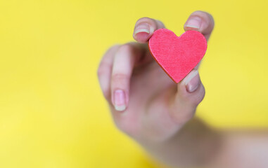 A woman's hand holding a red heart on a yellow background. selective focus. A wooden valentine in a blurry female hand. valentine's day. World Blood Donor Day, charity and care, blood donation