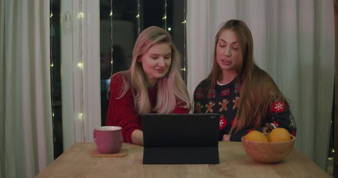 Young women at Xmas using tablet video call. Young girls laugh and talk. Christmas lights decorated with ornament pullovers in dining room at home.