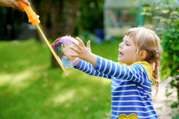 Happy little blonde preschool girl having fun with blowing soap bubble blower. Cute child playing...