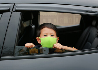 Asian little boy wearing hygiene face mask pokes his head out of car window during coronavirus...