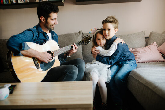 The father plays an acoustic guitar for the children at home.