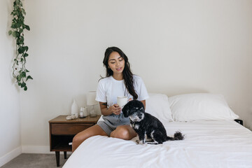 Young Filipino woman sitting on the bed with coffee and dog