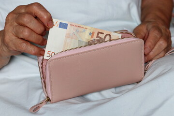 Elderly woman takes out a euro note from her wallet sitting in bed. Concept of pension payments,...