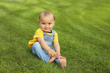 a beautiful little boy in a yellow T-shirt and a denim suit is sitting on the lawn in the park
