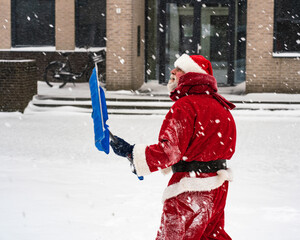 Joyful Santa Claus in a traditional costume with a shovel removes snow on a snowy street.