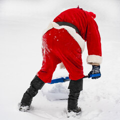Santa Claus in a traditional costume with a shovel removes snow on a snowy street. View from back.
