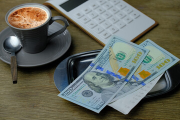 small gray cup of coffee on a wooden table, metal money tray, american dollars banknotes, Restaurant bill, calculator, tip money concept, home economy, saving on food