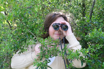 sly curious European adult woman holding black field binoculars with zoom in her hands, hiding in...