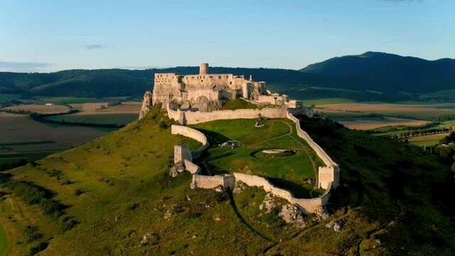 Aerial, scenic footage of Spiš Castle, lit by evening sun. Extensive castle ruins lit by orange light, against mountains in background. Famous tourist place, Slovakia.