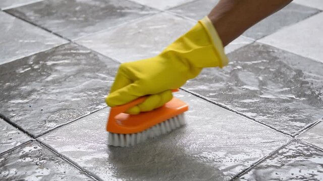 Hands wearing yellow rubber gloves are using a plastic floor scrubber to scrub the tile floor with a floor cleaner.