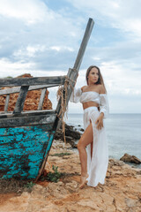 Sexy woman in a white dress at a wrecked ship on a rocky seashore