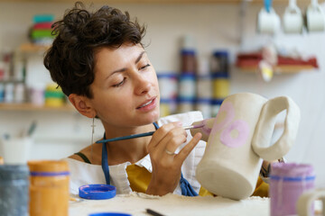 Concentrated woman potter holding paintbrush, painting on clay jug in workshop, working in pottery...